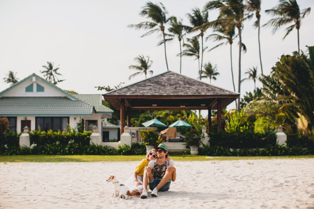 young stylish hipster couple in love walking playing dog puppy jack russell, tropical beach, cool outfit, romantic mood, having fun, sunny, man woman together, horizontal, vacation, house home villa